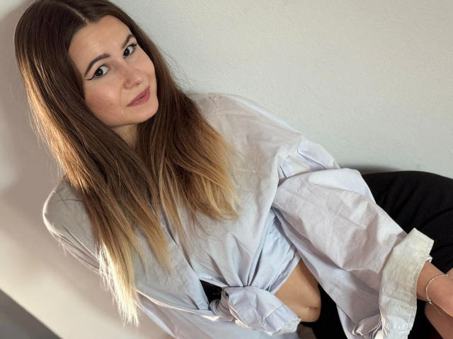 AliceeCute-hot with long brown hair, wearing a tied white shirt, black pants, and eyeliner, leaning against a wall, showing a subtle smile, with light in her hair.