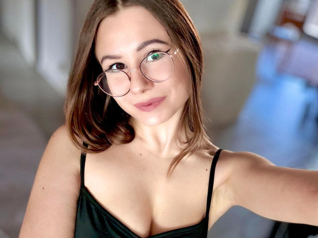AliceeCute-hot with green eyes, wearing round glasses and a black spaghetti strap top, showing her tits in a selfie. She has light skin and brown hair.