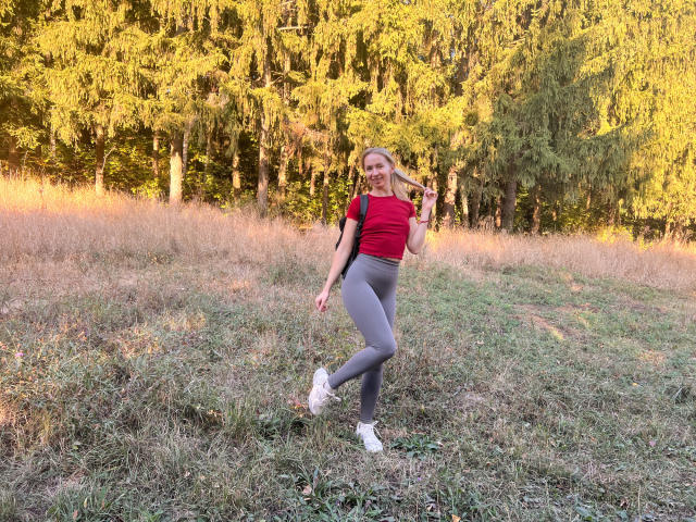 VasilisaSexy with blonde hair, red top, gray leggings, white sneakers, standing in grassy field, holding a backpack, smiling, sunlight, forest background.