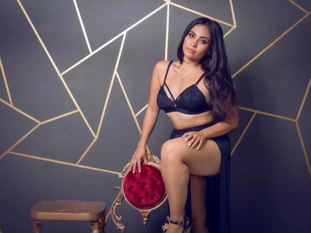 AliciaBecker-hot, dark-haired, wearing black lace bra and sheer skirt, sits on red velvet chair with gold trim, one leg up, showing her ass, against geometric gold lines.