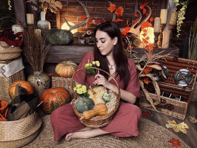 KaleyReed with long brown hair, red dress, holding wicker basket with pumpkin, apple, yellow flowers, sitting on straw, surrounded by pumpkins, candles, autumn leaves.