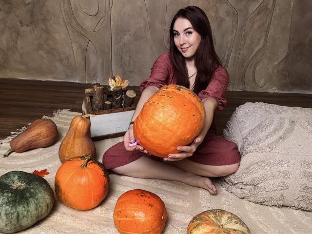 KaleyReed, with long brown hair, sits on a beige rug holding a huge orange pumpkin. She's wearing a red dress, surrounded by various pumpkins and a butternut squash.