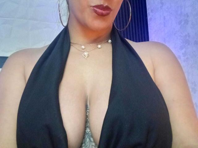 DayanaMarians69, avec de grandes seins, porte un haut noir à enfiler qui révèle une profonde décolleté, un collier en or et des boucles d'oreilles cerceaux. Elle a un rouge à lèvres rouge foncé et un bronzage léger..