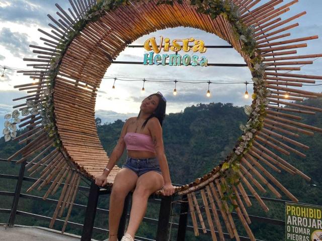 Mafert with small tits, wearing a pink strapless top and denim shorts, sits on a wooden frame with a sunburst design, looking up with a smile.