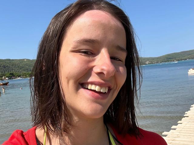 LenaBlush-hot hat feuchtes, braunes Haar, ein rotes Hemd und ein fröhliches Lächeln, das weiße Zähne zeigt. Sie ist an einem Strand mit klarem, blauem Wasser und einem hölzernen Steg..