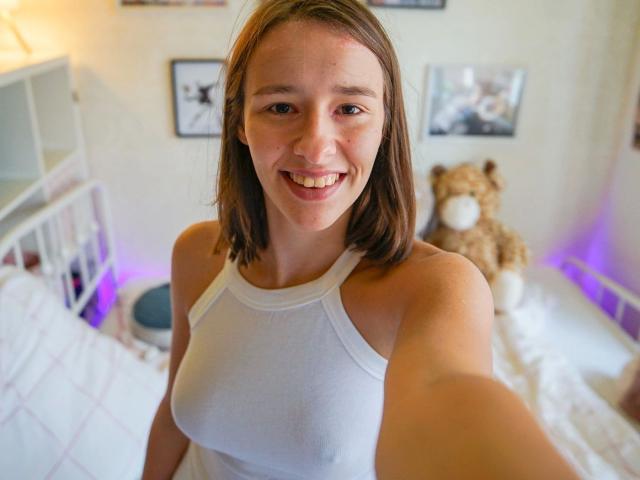 LenaBlush – heiß, mit braunen Haaren, lächelnd, trägt ein weißes Tanktop, zeigt kleine Brüste, macht ein Selfie in einem hellen Raum, im Hintergrund ist ein Teddybär zu sehen..