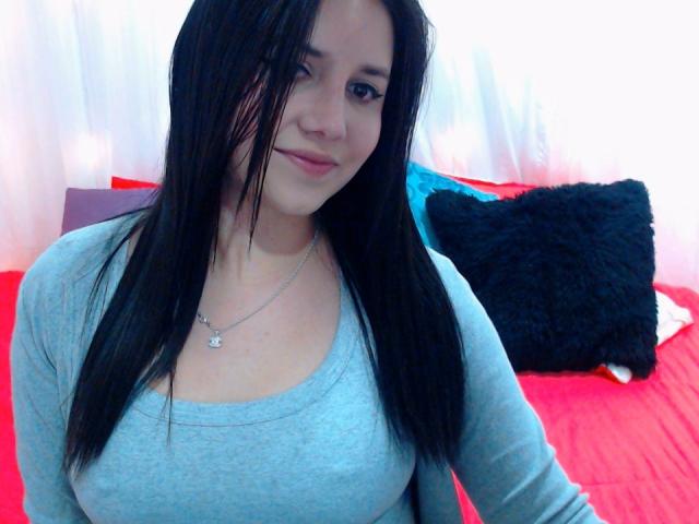 Kkatalina with long black hair, light skin, small tits, wearing a grey shirt and necklace, sitting on a red bed with a black pillow.