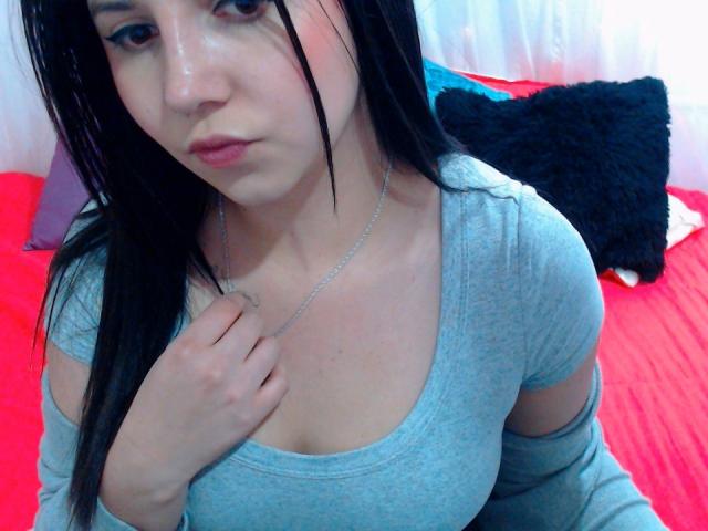 Kkatalina with long black hair, wearing a grey off-shoulder top, has her hand on her chest, showing her tits. She has a silver necklace and a serious expression.