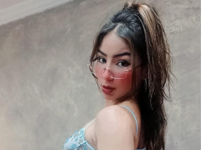 AshleyMiler with wet brown hair in a ponytail, wearing pink glasses and a blue lace top, showing off her tits and a sexy, confident look.