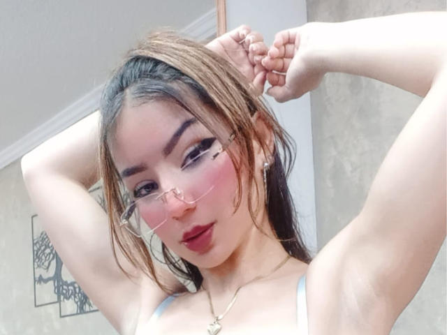 AshleyMiler with arms raised, wearing pink glasses, red lipstick, and a white top, showing her armpits and small tits, has brown hair and a gold necklace.