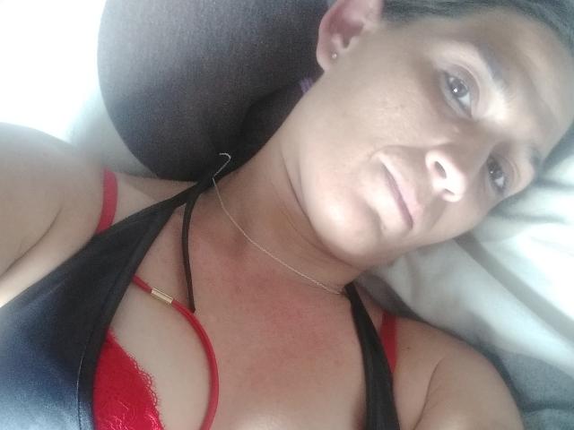 HotSexyLady69 with light brown skin, black headband, red lace bra, black halter top, and necklace, lying down, smiling slightly, showing her tits and a subtle tan line.