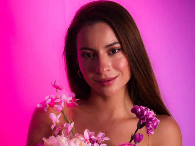 Daphne Blakee, met roze-paarse bloemen in haar haar, lacht. Ze heeft lang bruin haar, een schattig gezicht, een kleine neus, roze lippenstift en draagt een top zonder mouwen. De achtergrond is roze en paars..