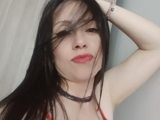 MondBlue with black hair, red lipstick, and a sparkly choker, raising her arm, showing a bracelet, and giving a slight smirk, with her hair in her face.