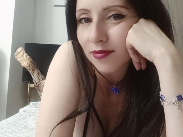 MondBlue with long black hair, pink lipstick, and blue butterfly necklace, laying on bed, head resting on hand, fishnet leg visible, wearing black lingerie, purple bracelet.