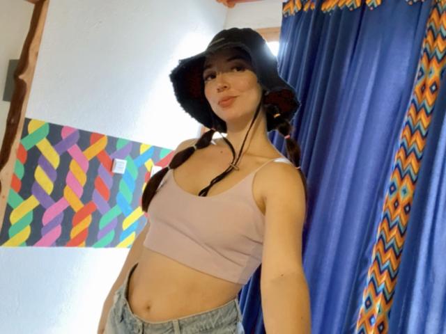 ValeryLombardi, con un sombrero negro, un top corto rosa y jeans azules, tiene senos prominentes, una nariz linda y trenzas largas, y está de pie frente a cortinas azules..