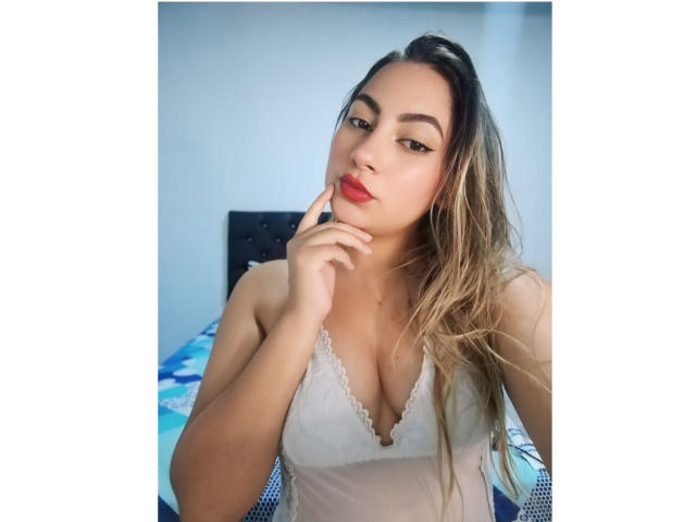 CamilaTeffey with long blonde hair, wearing a white lace top, touches her red lipstick with her finger, showing her tits and a sexy, confident expression.