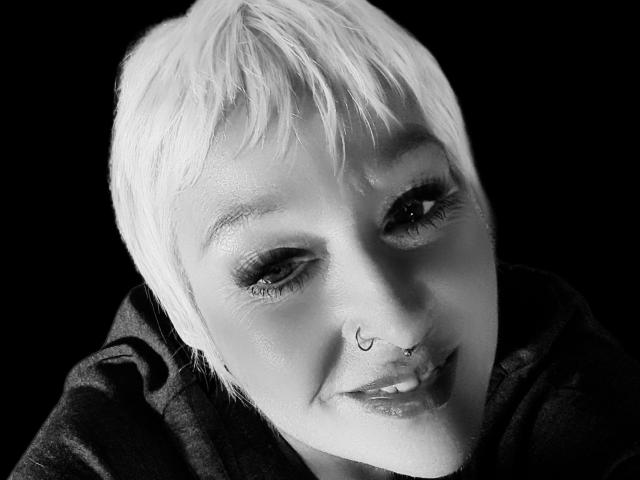 Black-and-white photo of NaughtyWillow with short, white hair, nose ring, and smiling, showing eyelashes, slight wrinkles, and wearing a dark hoodie against a black background.