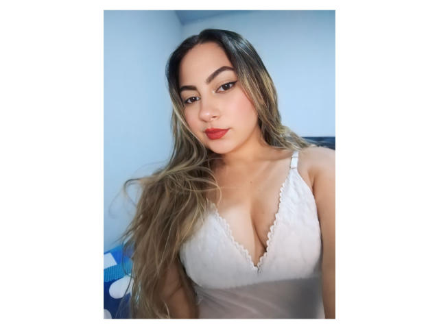 CamilaTeffey with long blonde hair, wearing a white lace bra, showing her big tits, red lipstick, and a slight smile, in a bright, blue-walled room.