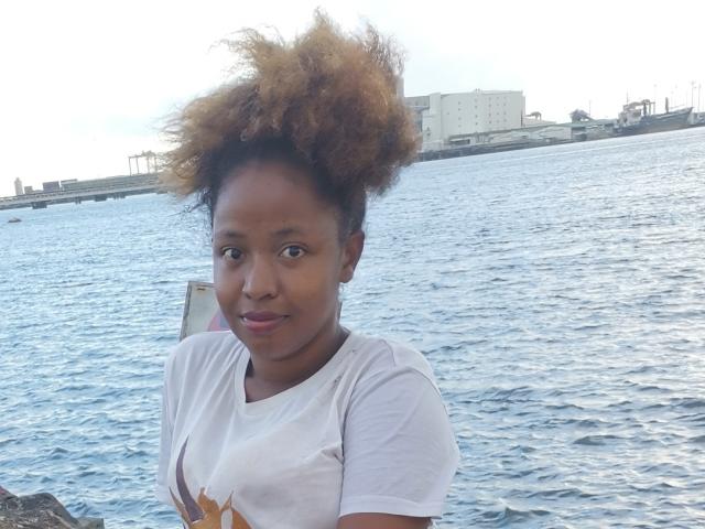 Michbelle, avec ses cheveux afro bruns foncés et bouclés, porte un t-shirt blanc et se tient près d'une étendue d'eau bleue, souriant légèrement et regardant directement la caméra. Ses seins sont petits et ses fesses ne sont pas visibles..