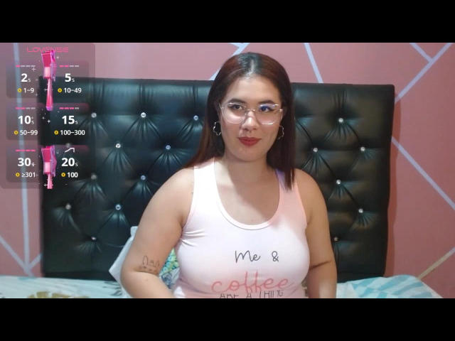 SophieMoonroe has brown hair, glasses, and a pink tank top showing her big tits. She's sitting on a bed with a black tufted headboard. Her tattoo is on her right arm.