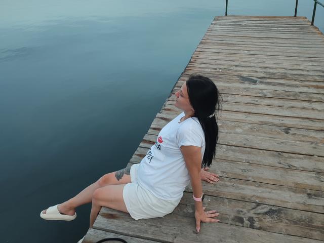 XCrazyBabeX assise sur un quai en bois, portant une chemise blanche et un short, des chaussures blanches, les cheveux noirs attachés en queue de cheval, un tatouage sur la cuisse gauche, regardant l'eau calme..