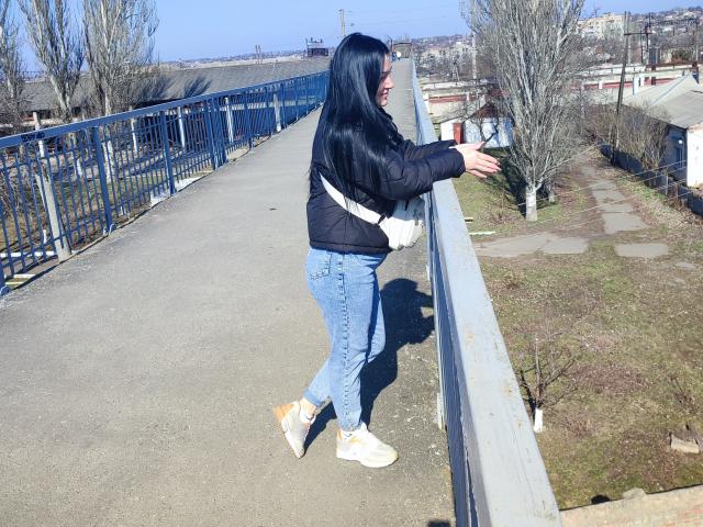 XCrazyBabeX con cabello negro largo, chaqueta negra, jeans azules, botas blancas, apoyada en una barandilla azul, mirando de lado, de pie en un camino de concreto, de día, cielo azul despejado..