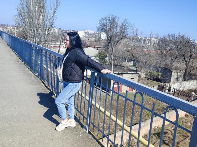 XCrazyBabeX has black hair, wearing a black jacket, blue jeans, and white sneakers, leaning on a blue metal railing with her right hand, on a sunny day.