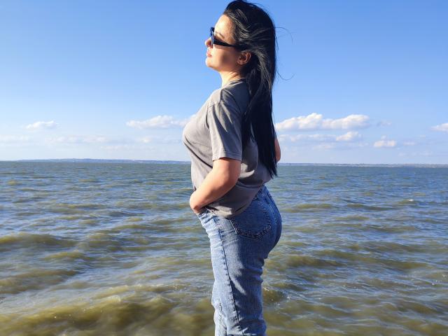 XCrazyBabeX standing in shallow water, wearing a gray t-shirt, blue jeans, and black sunglasses, with hands in pockets, showing a curvy ass and long black hair.