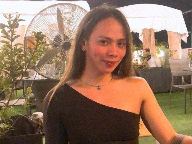SuckableMistress - sensual, com longos cabelos castanhos, blusa preta de um ombro só, seios pequenos, colar, sorriso discreto, em pé em frente a uma grande ventoinha, noite, ambiente externo..