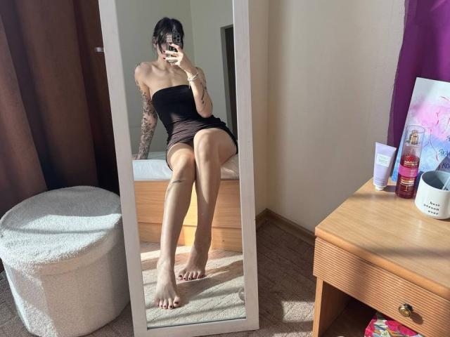 Tsukerberg, in a black strapless dress, sits on a bed, showing bare feet, tattooed arm, and legs in a mirror selfie. Her ass is visible. Mirror, bed, and wooden side table.