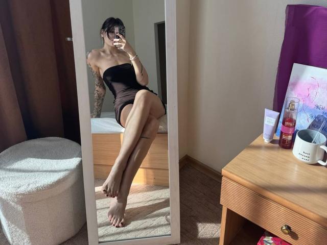 Tsukerberg in a black strapless dress, showing off her bare ass and tattooed arm, taking a mirror selfie with her phone, legs crossed, on a wooden floor.