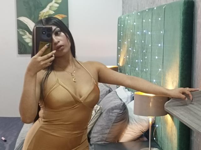 EmmaaRodriguez in a tight, tan, halter dress, showing her big tits and curvy ass, taking a mirror selfie with her right hand on a green headboard.