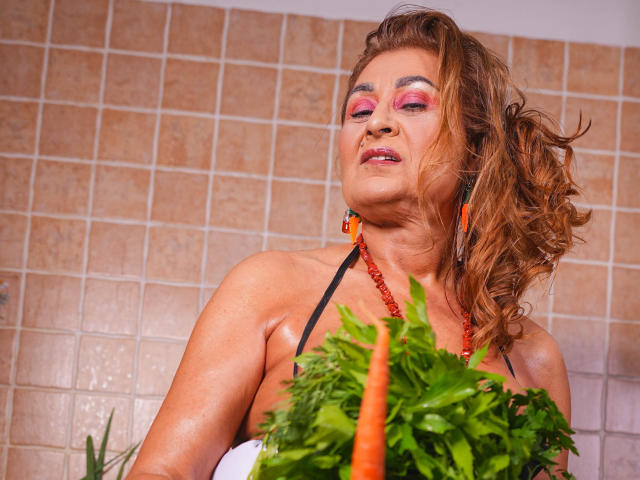 Karla Hartley, con i capelli castano ondulati, ombretto rosa e perline rosse, tiene in mano carote fresche e prezzemolo, guarda verso l'alto con le labbra leggermente dischiuse, indossando un top nero con spalline..