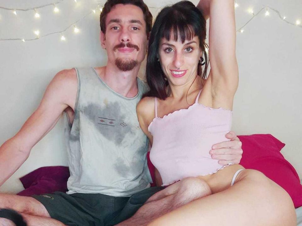 AngelaAndHector with small tits in a pink tank top, and Hector with a dirty gray tank top and visible armpit hair, sitting on a bed, arms around each other.