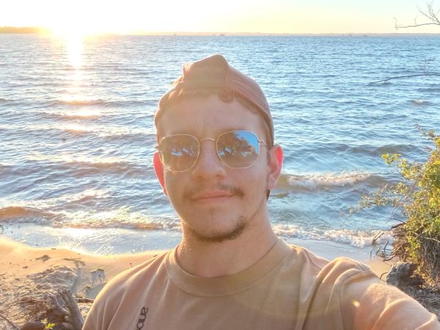 FrenchBibi with a beard, wearing a brown cap and reflective aviator sunglasses, smiling at the sunlit beach, reflecting water in his lenses, brown shirt, and a mustache.