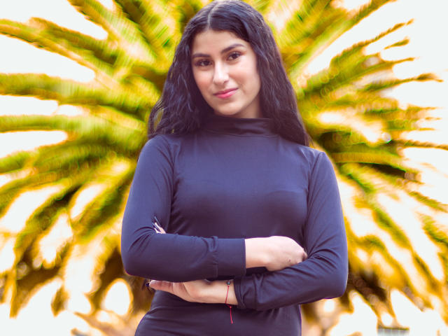 EmaHenderson with long black hair, wearing a black long-sleeve top, arms crossed, standing in front of a green palm tree, smiling slightly, pink lipstick, red bracelet.
