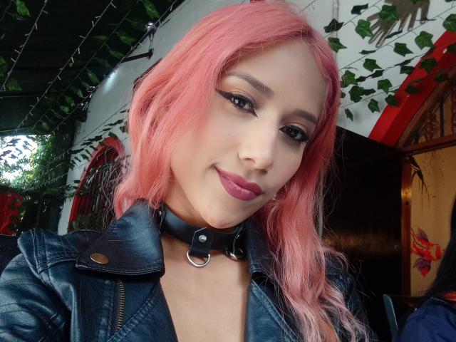 BrianaHarpers with pink hair, black leather choker, and denim jacket, showing off her pink lipstick and eyeliner. Her tits are small and she's smiling at the camera.