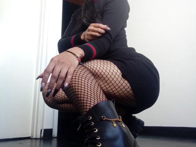 BrianaHarpers squatting in black rib-knit dress, fishnet stockings, and black lace-up boots with gold laces. Her hand is on her knee, showing long, dark nails.