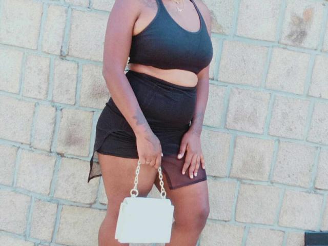 Jobba with dark skin, wearing a black sports bra and skirt, holding a white chain purse, showing her ass and tits, standing against a brick wall.