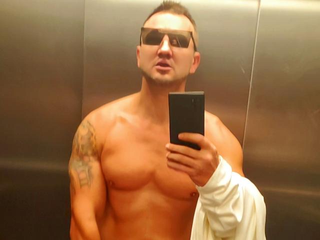 DestinyChris, sin camisa, musculoso, con un tatuaje en el hombro derecho, usando gafas de sol, sosteniendo un teléfono, con expresión de labios fruncidos, con una toalla blanca en la mano izquierda, tomando una selfie en un ascensor, con cabello corto castaño, de piel media..