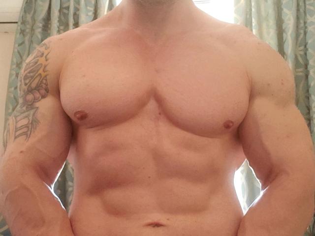 DestinyChris, sin camisa, musculoso, con abdominales y pectorales definidos, tiene un tatuaje en el brazo derecho y piel pálida. Los pezones de DestinyChris son visibles, y está de pie frente a unas cortinas..