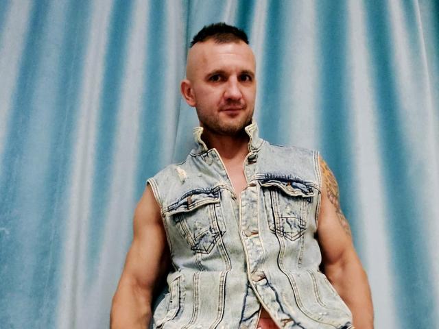 DestinyChris es un hombre musculoso con la cabeza rapada y un tatuaje en su brazo derecho, que lleva un chaleco vaquero con un brazo desgarrado, y está de pie frente a una cortina azul..