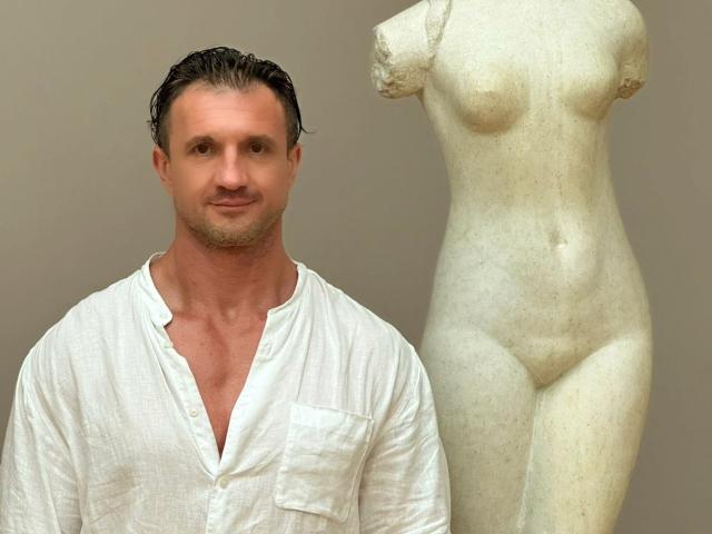 DestinyChris, torse nu, portant une chemise blanche à boutons, se tient à côté d'une statue en marbre blanc représentant une femme avec des seins, des fesses et un vagin. DestinyChris a les cheveux mouillés et plaqués en arrière, un léger sourire et un bronzage..