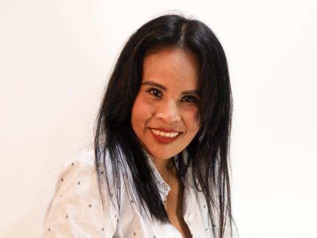 Anthoanela with medium brown skin, black straight hair, smiling, wearing a white button-up shirt with small black dots, looking at the camera, showing her teeth.