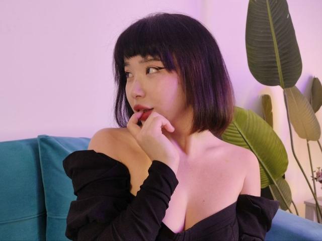 EveLovelace avec une coupe de cheveux courte noire, des lèvres pulpeuses, un doigt dans la bouche, un haut noir à épaules dénudées, montrant ses seins, une plante verte sur la droite, un canapé bleu, un éclairage violet..