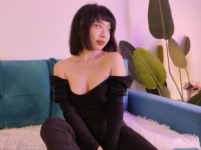 EveLovelace com cabelo preto curto, blusa preta com um ombro descoberto, mostrando os seios, sentada em um sofá azul, tapete branco felpudo, plantas verdes ao fundo, olhando para o lado, iluminação rosa..