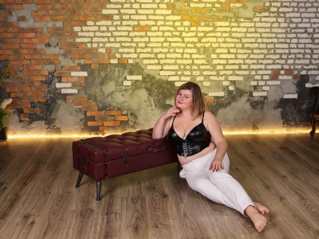 ZannaDreil with medium tits, wearing a black leather bra, white pants, and barefoot, sitting on a red leather bench, wood floor, brick wall, hand on chin, smiling.