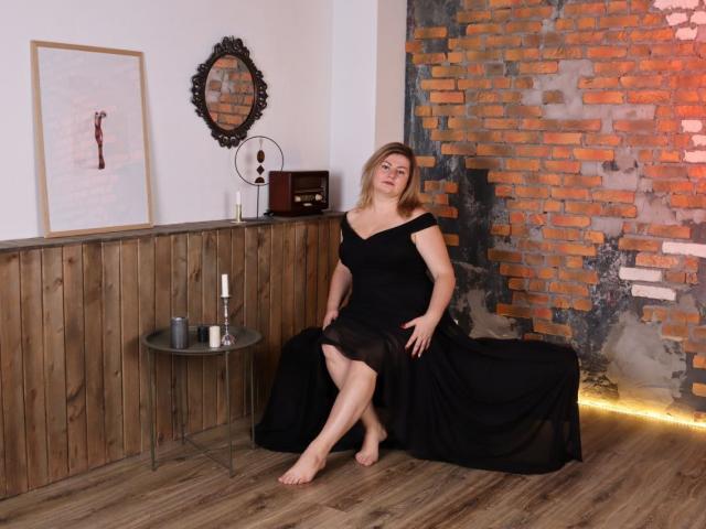 ZannaDreil with bare feet, black off-shoulder dress, sitting on wooden chair, holding dress hem, wooden panel wall, brick wall, round table with wine bottle, mirror, and clock.