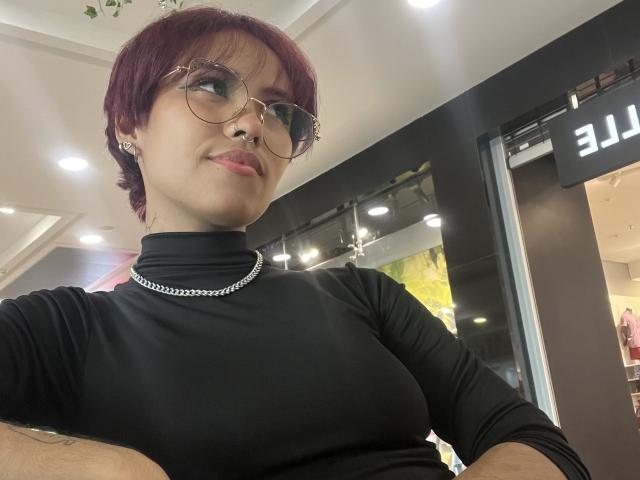 RoussPark with short red hair, glasses, black turtleneck, silver chain, and arms crossed, showing small tits, smiling, in a well-lit indoor space.