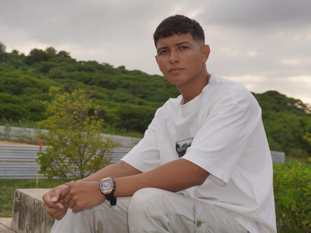 YeiSant, con cabello castaño corto, ojos marrones y piel morena, vestida con una camisa y pantalones blancos, sentada con las manos en las rodillas, un reloj negro en la muñeca, y un bosque verde en el fondo..
